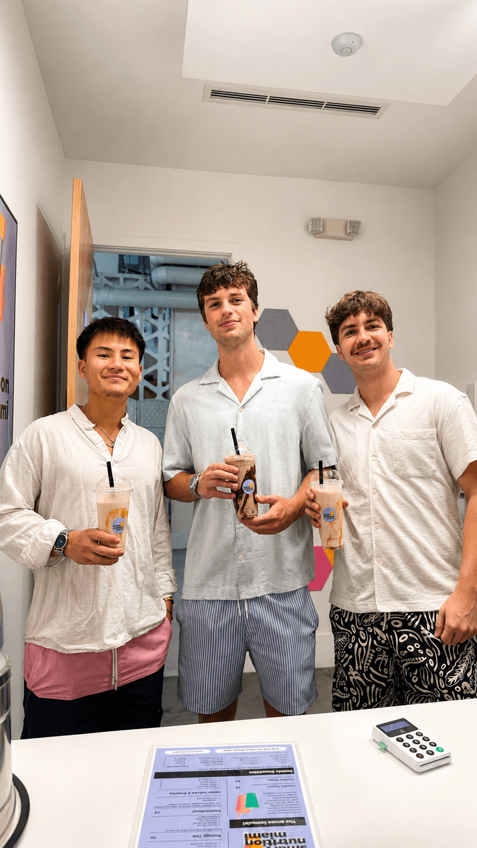 Three young men smiling and holding smoothies behind a counter in a bright cafe.