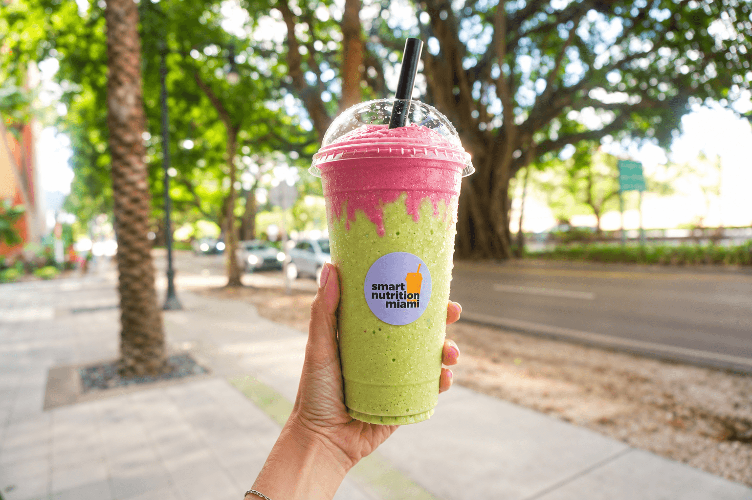 Hand holding a layered green and pink smoothie from Smart Nutrition Miami outdoors.