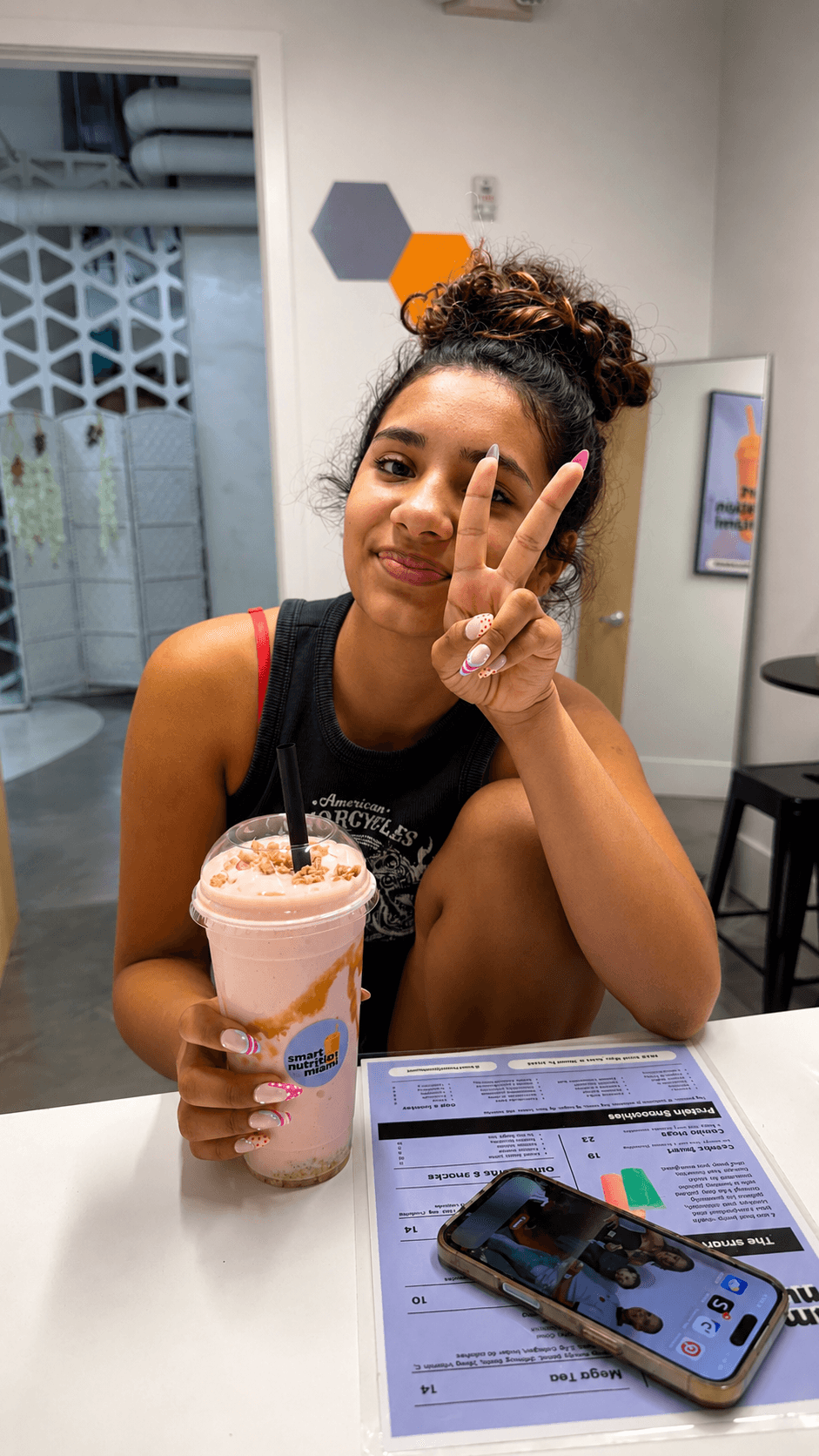 Young woman with curly hair holding a pink smoothie and making a peace sign.