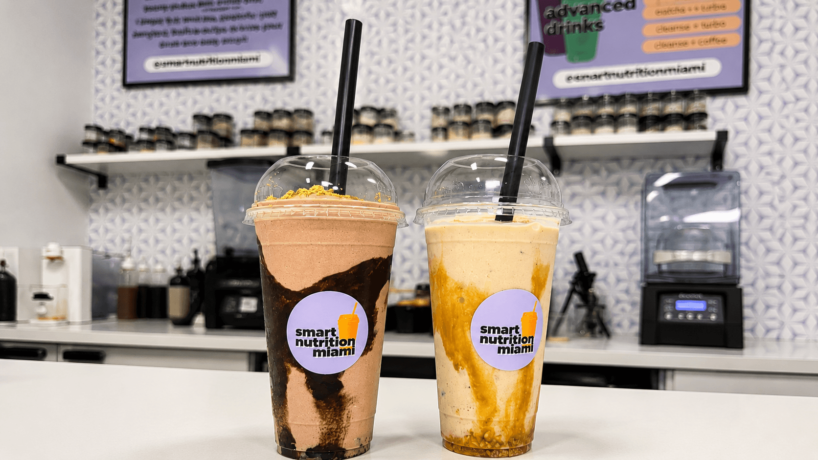 Two large smoothies with chocolate and caramel swirls in branded cups at a juice bar.