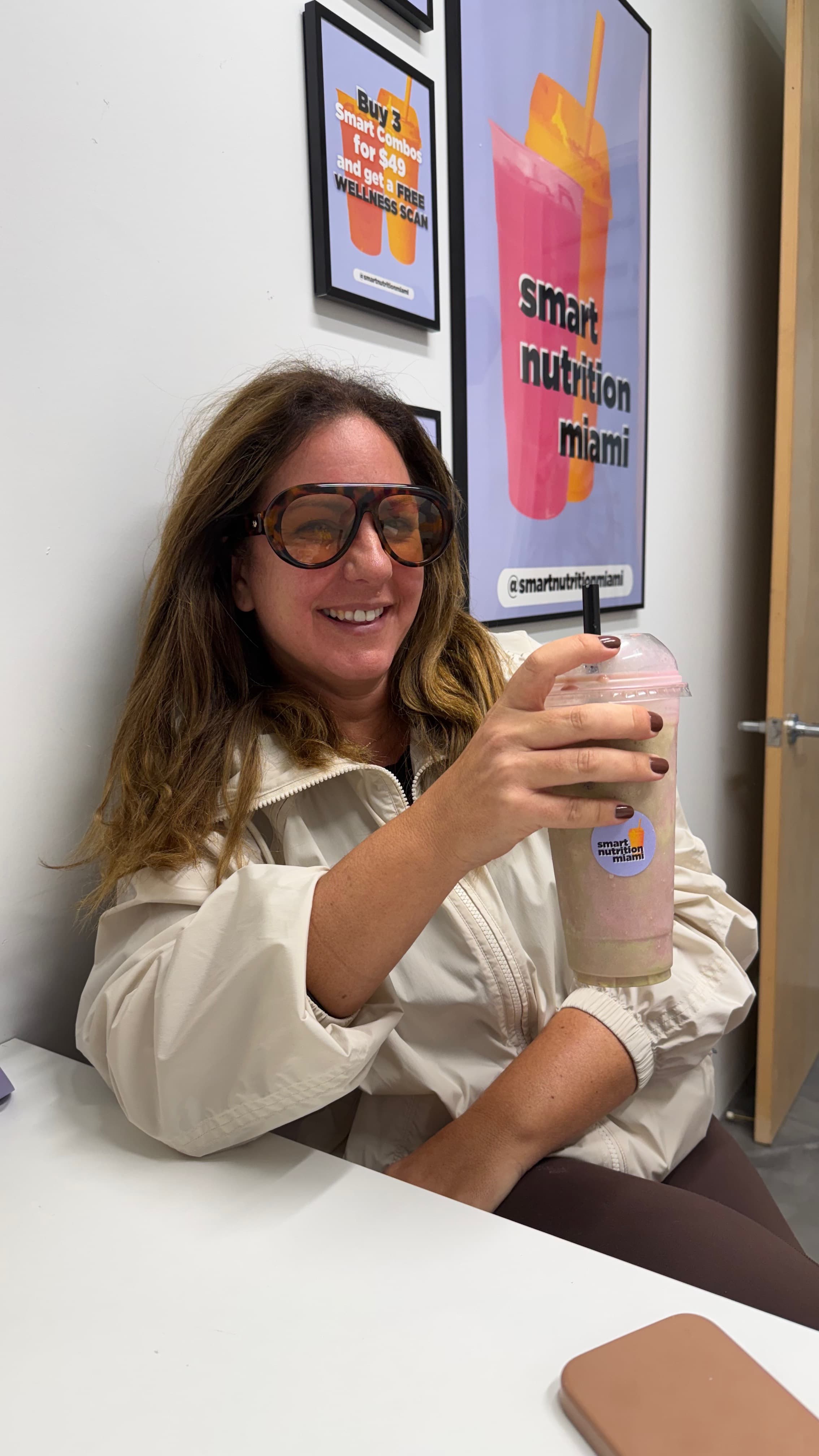Smiling woman wearing large sunglasses holds a smoothie in front of Smart Nutrition Miami posters.