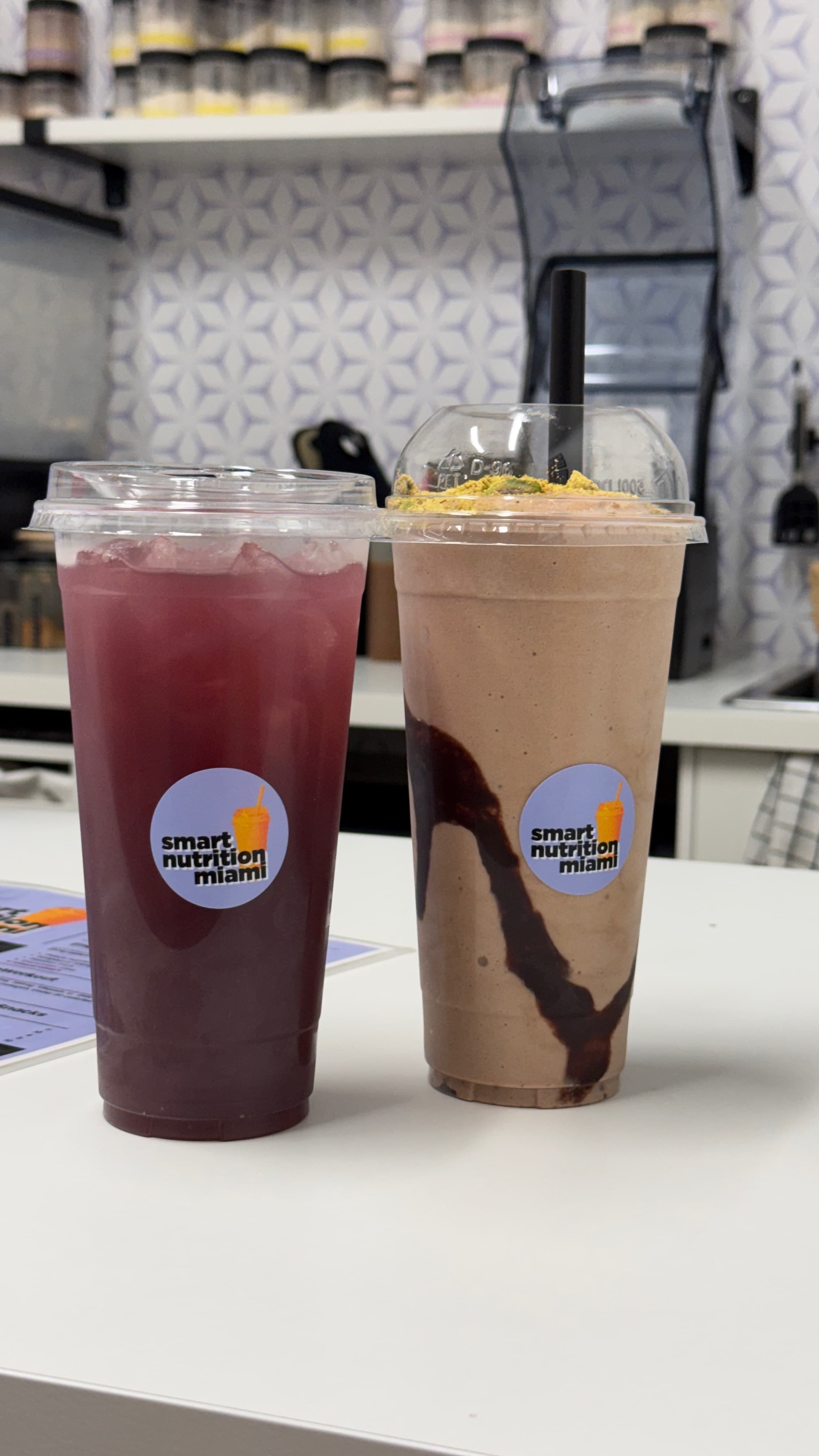 Purple iced beverage and chocolate smoothie with yellow toppings from Smart Nutrition Miami on counter.