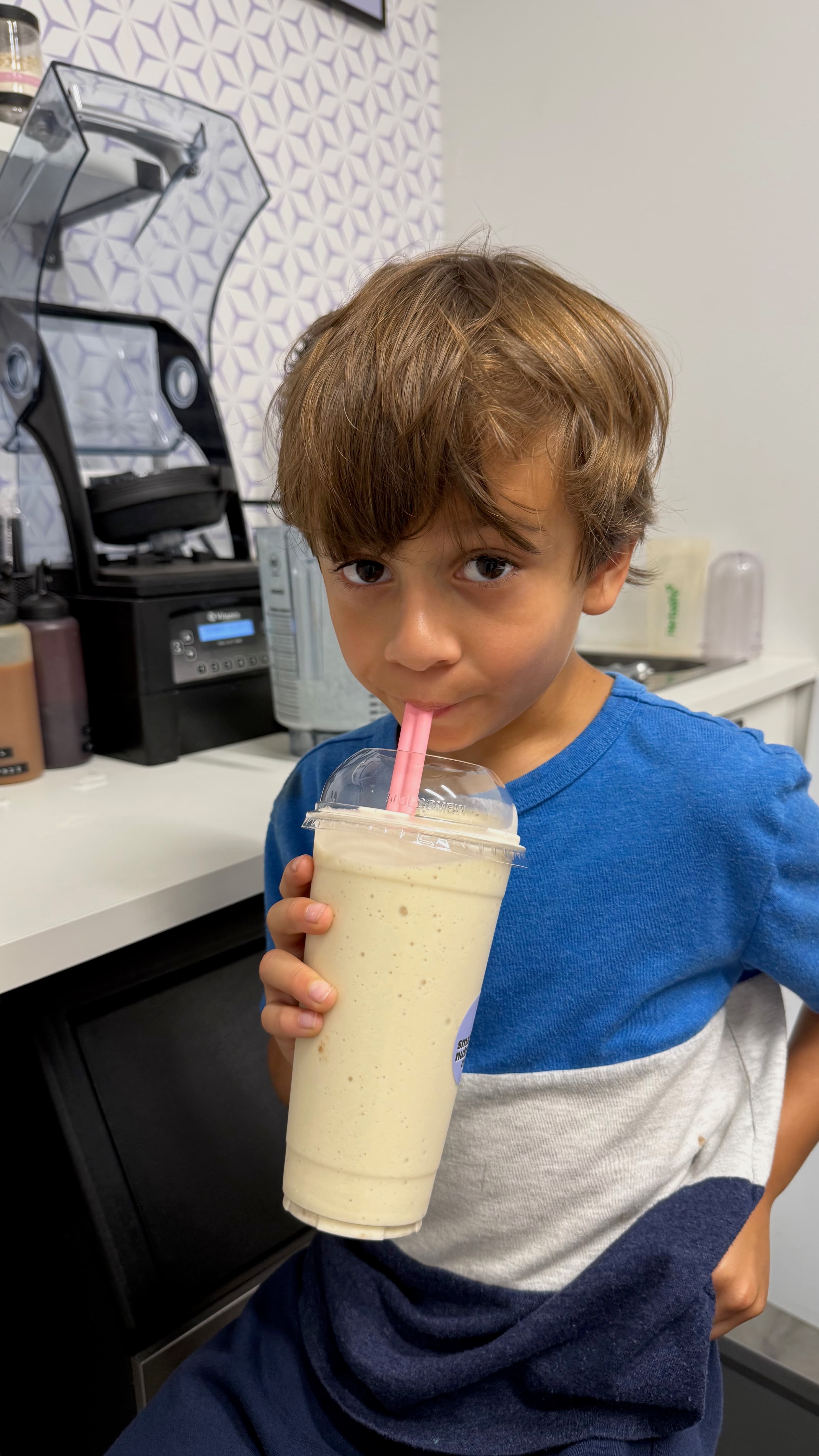 Young boy in a blue shirt sips a creamy smoothie through a pink straw.