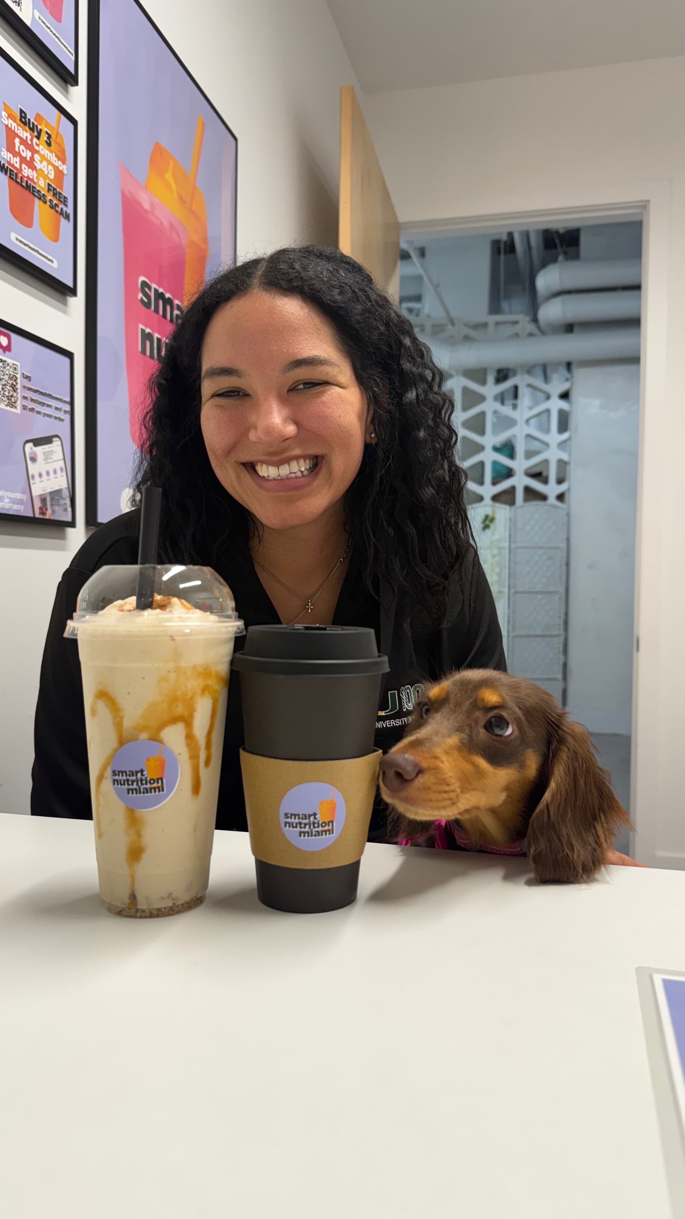 Smiling woman and dachshund dog sit behind a smoothie and coffee cup on a table.