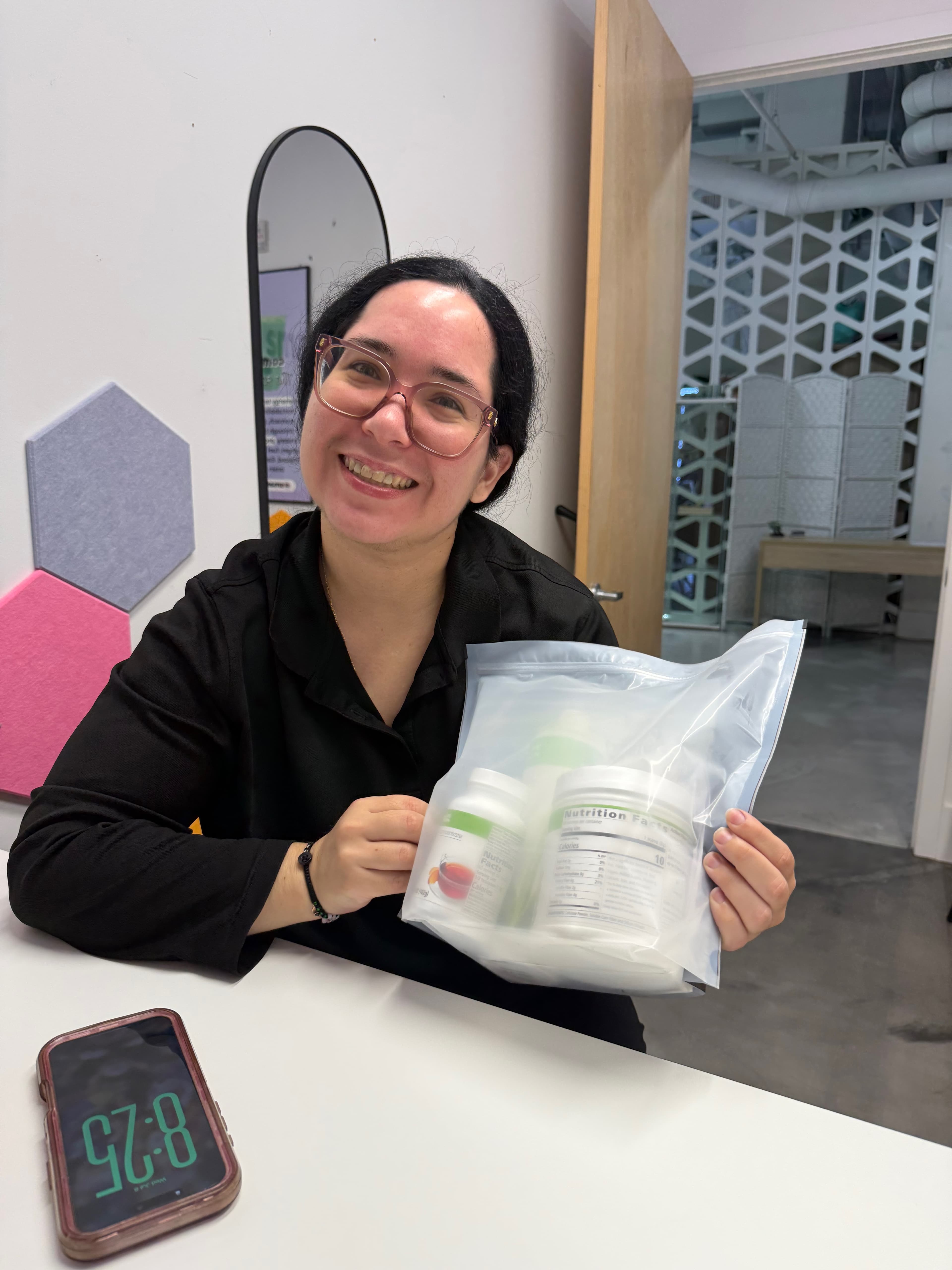 Smiling woman wearing pink glasses holds a clear bag filled with white nutritional supplement containers.