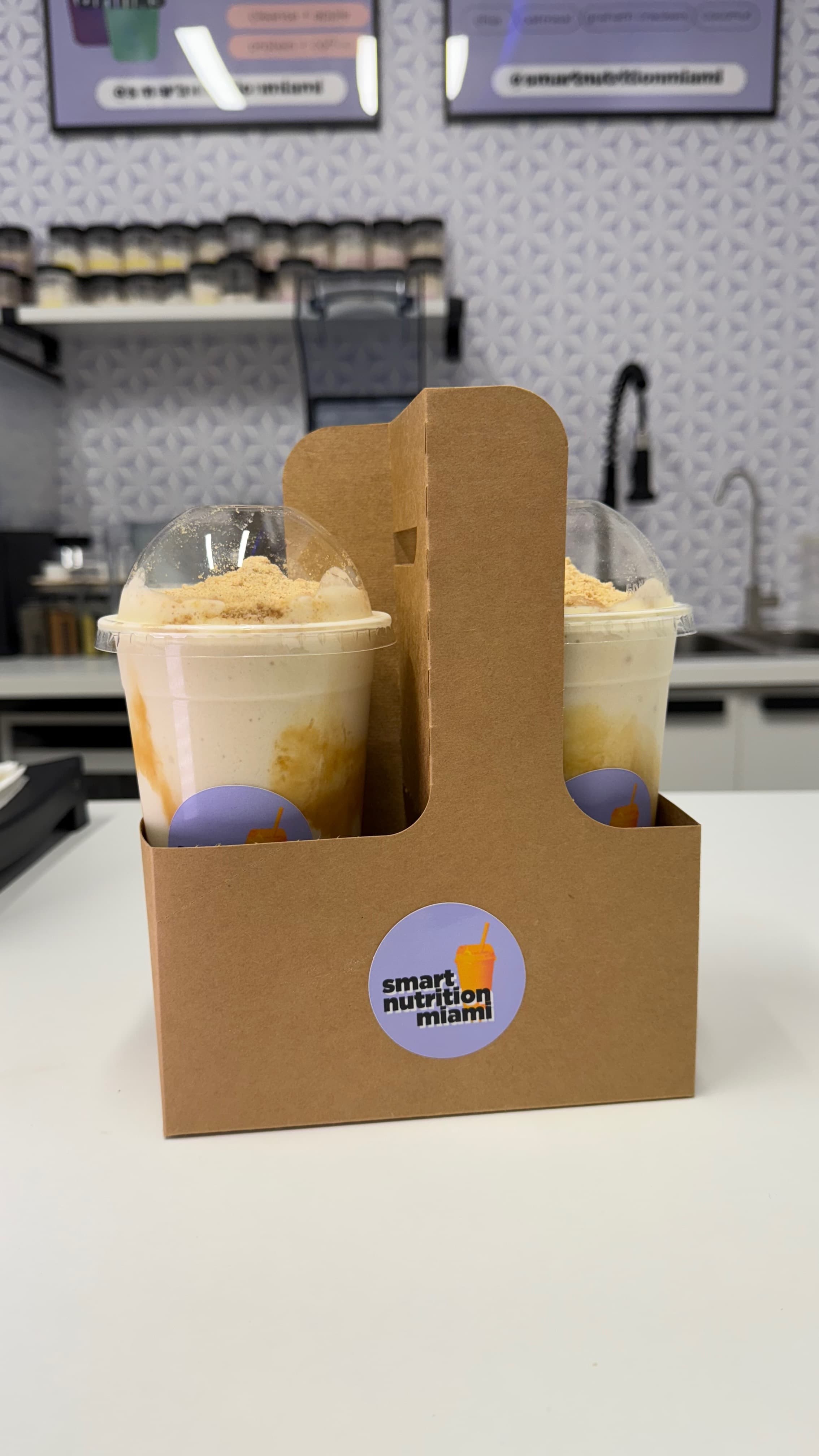 Two creamy smoothies with caramel swirls and crumble topping in a brown cardboard drink carrier.