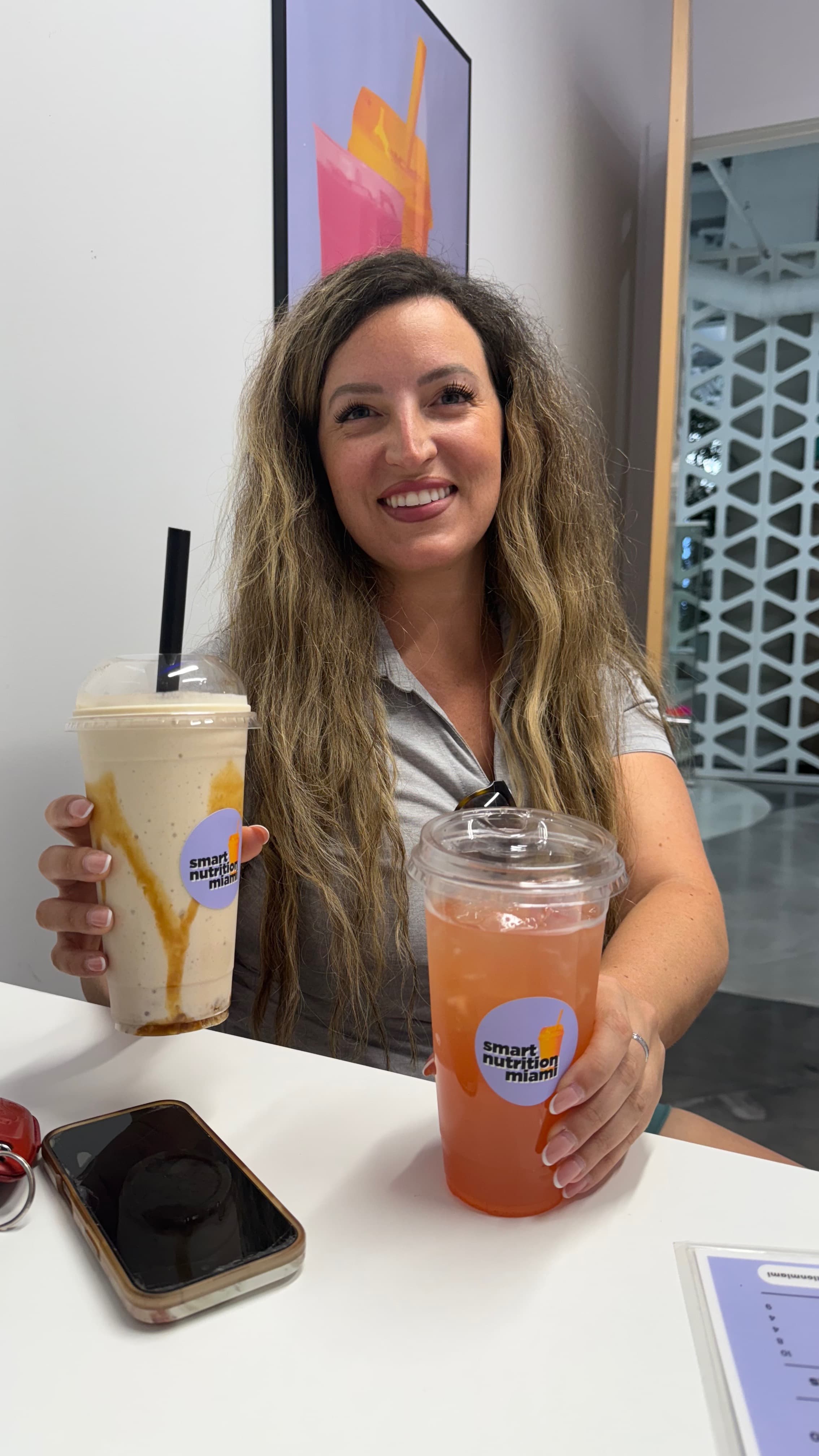Smiling woman with long wavy hair holds a caramel-drizzled shake and a pink drink.