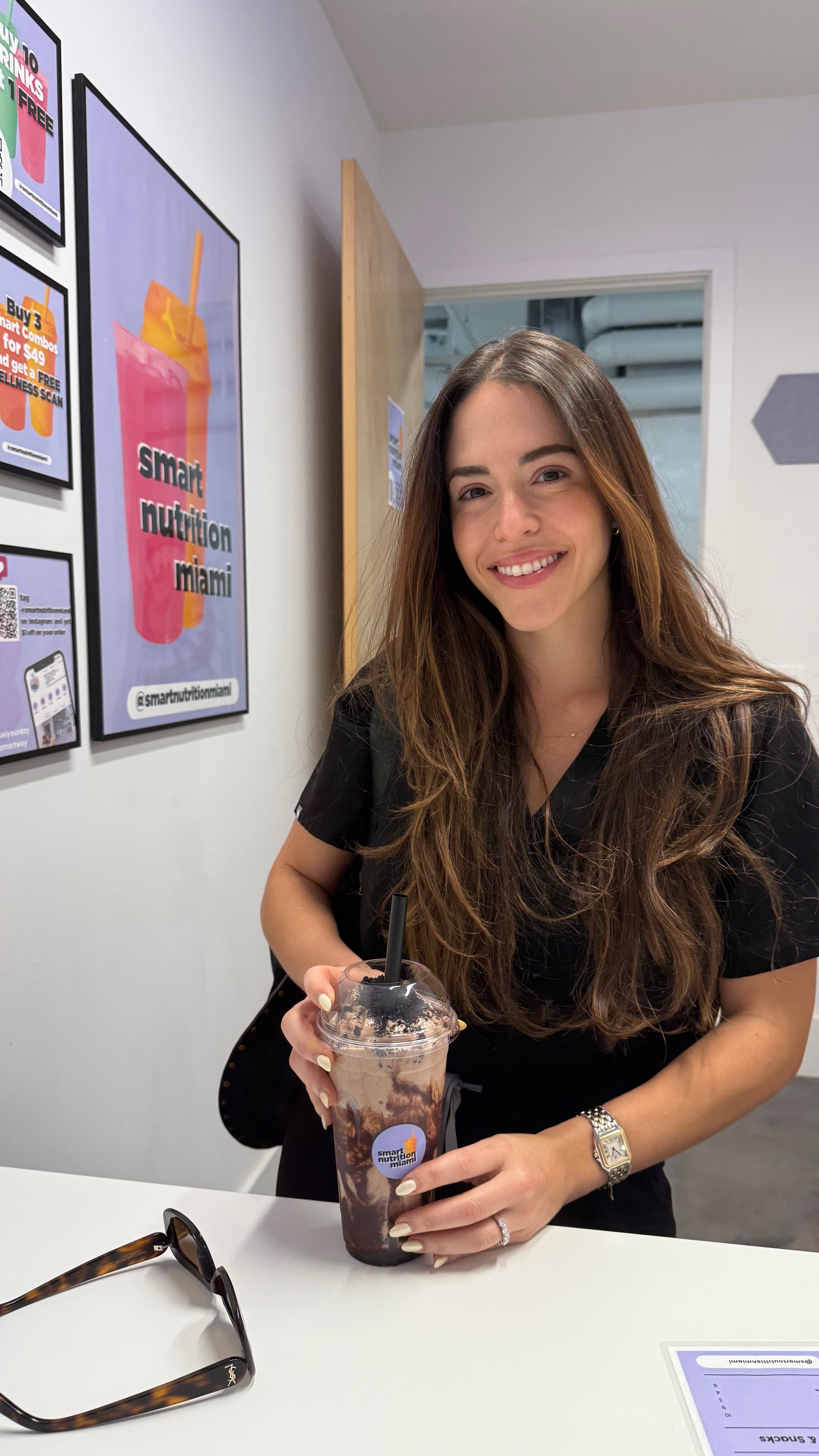 Smiling woman with long brown hair holds a chocolate smoothie at a nutrition shop counter.