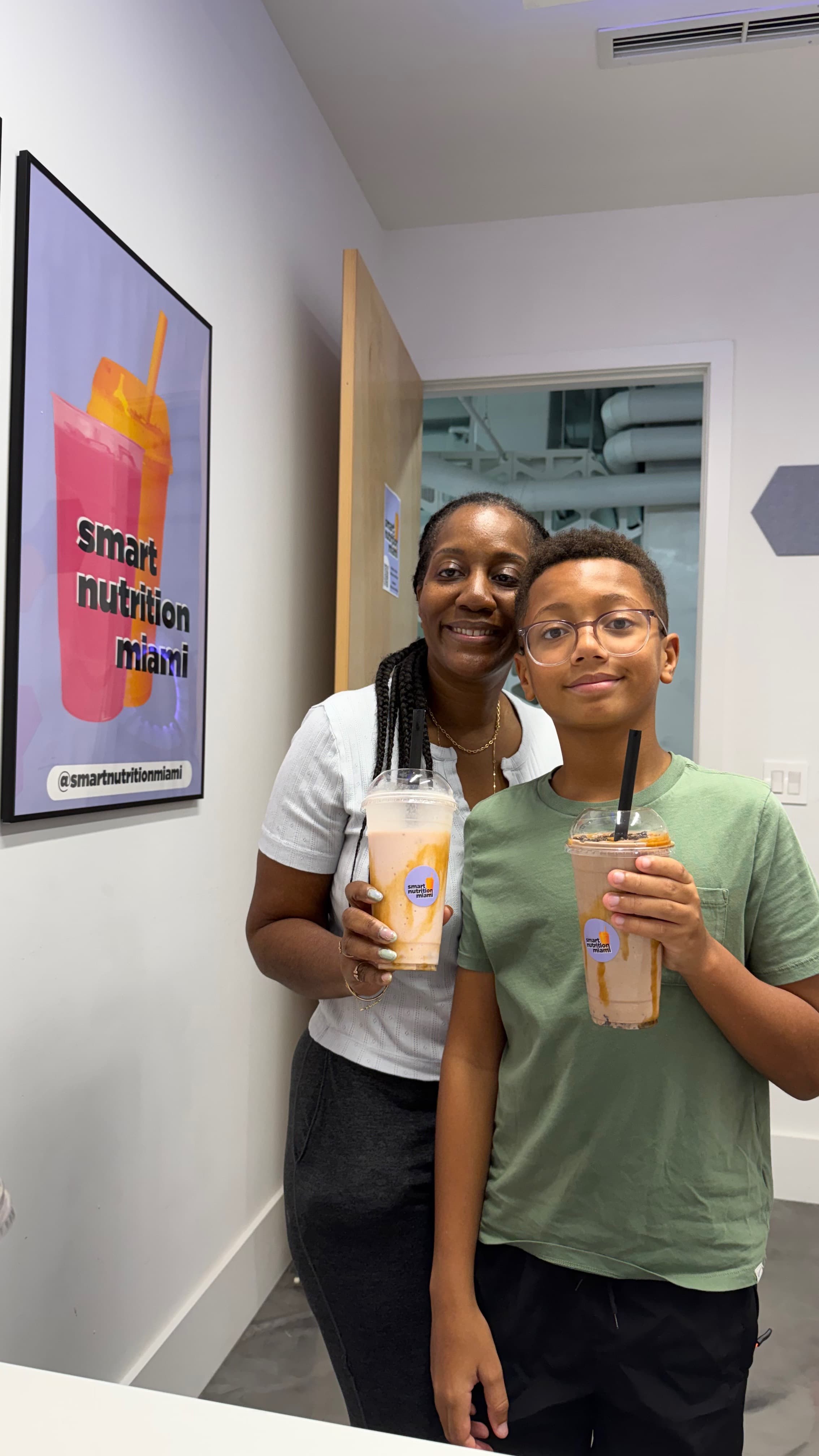 Smiling woman and boy holding smoothies in front of a Smart Nutrition Miami wall poster.