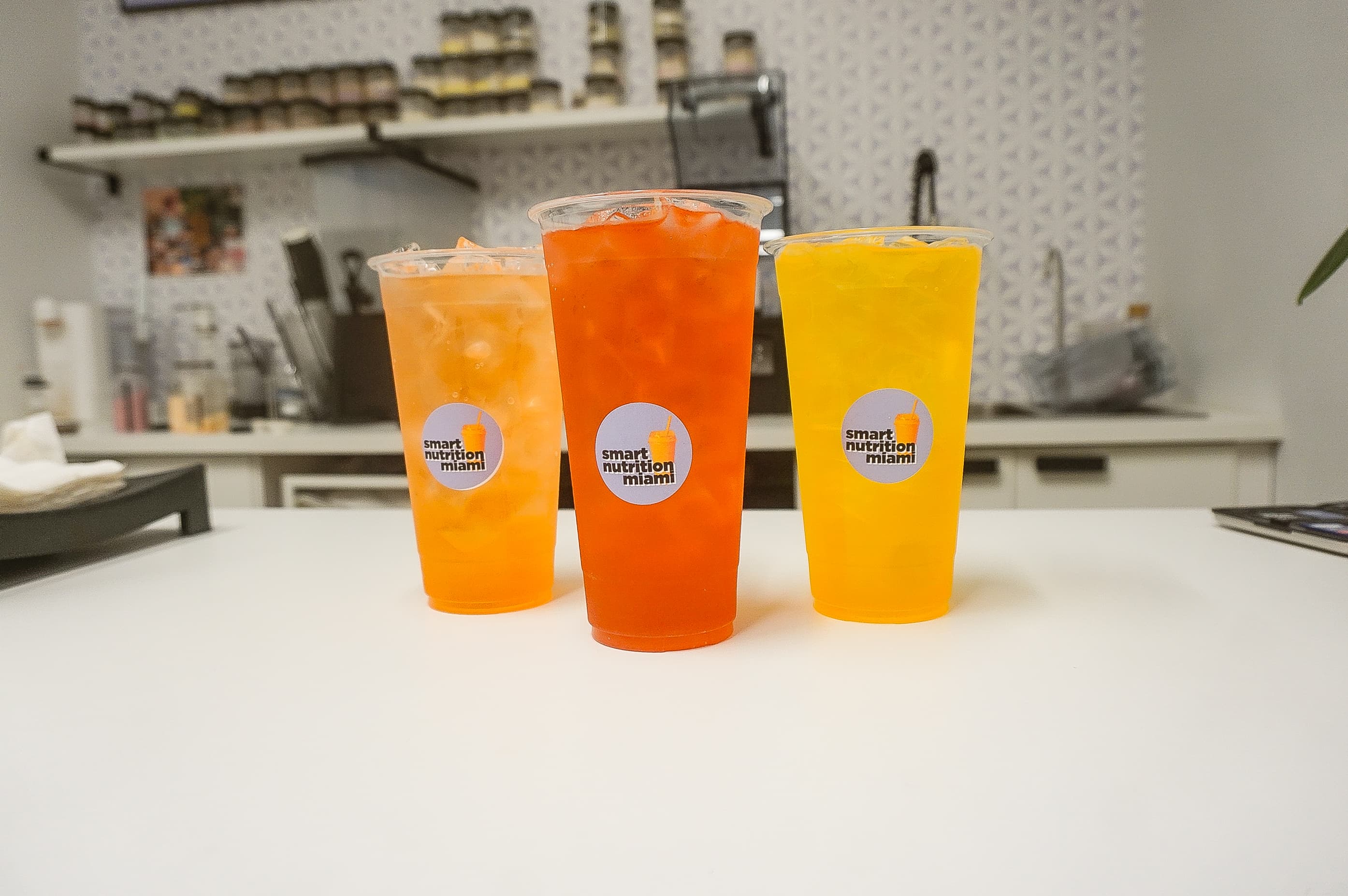 Three vibrant orange and yellow iced drinks with brand labels on a clean white counter.