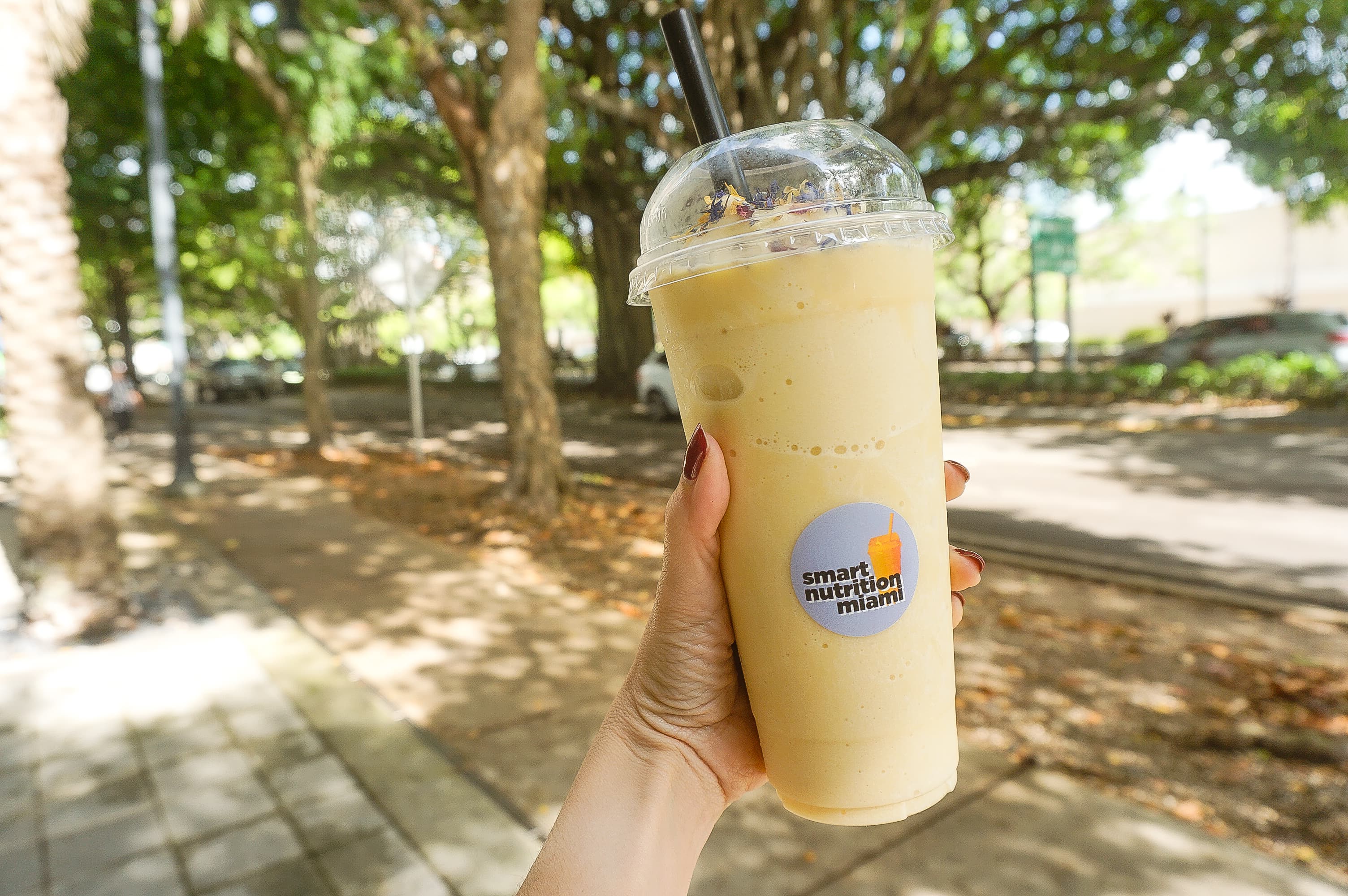 Hand holding a yellow smoothie with a logo sticker on a sunny, tree-lined street.