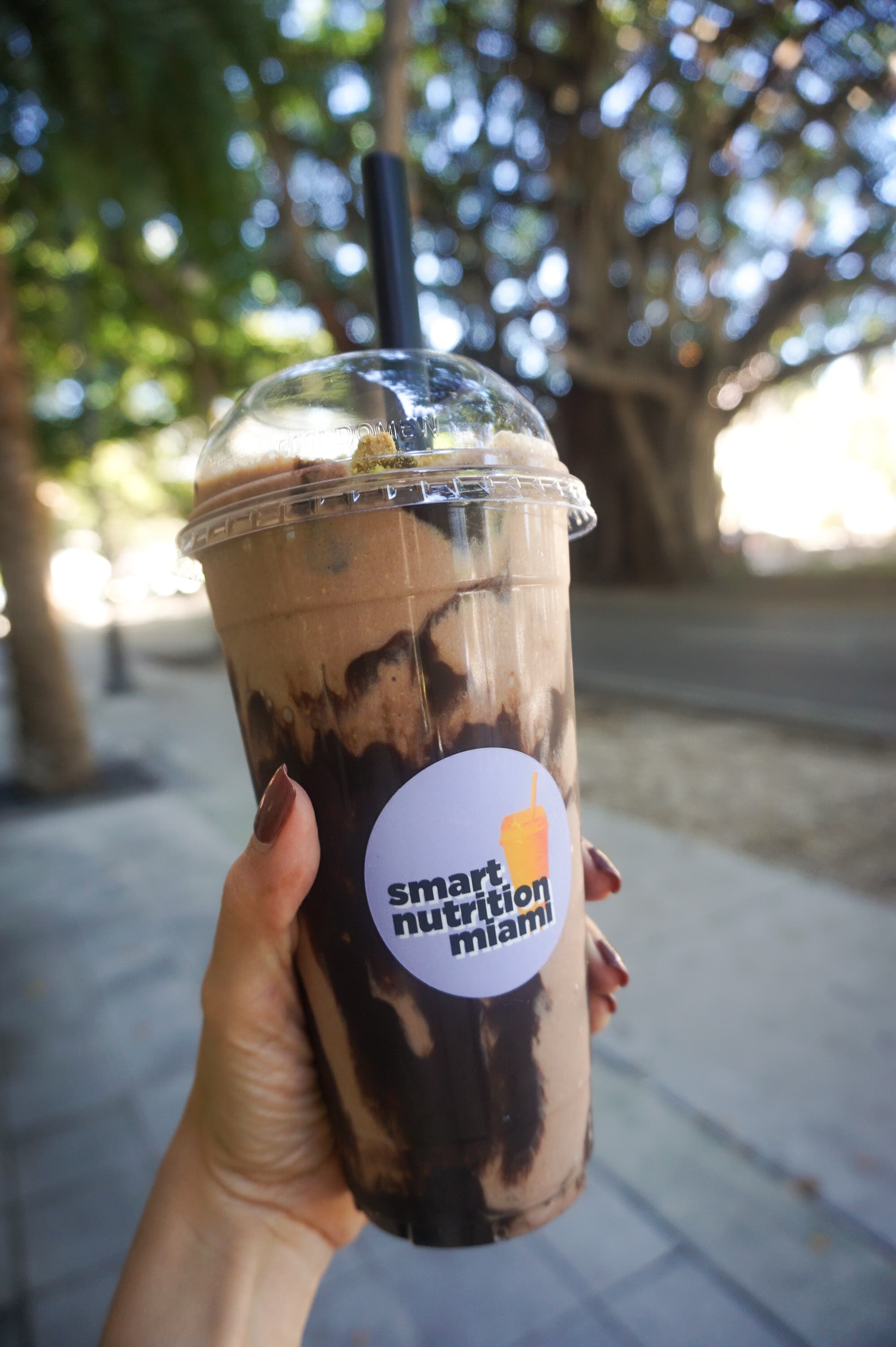Hand holding a chocolate smoothie with dark syrup swirls from Smart Nutrition Miami outdoors.