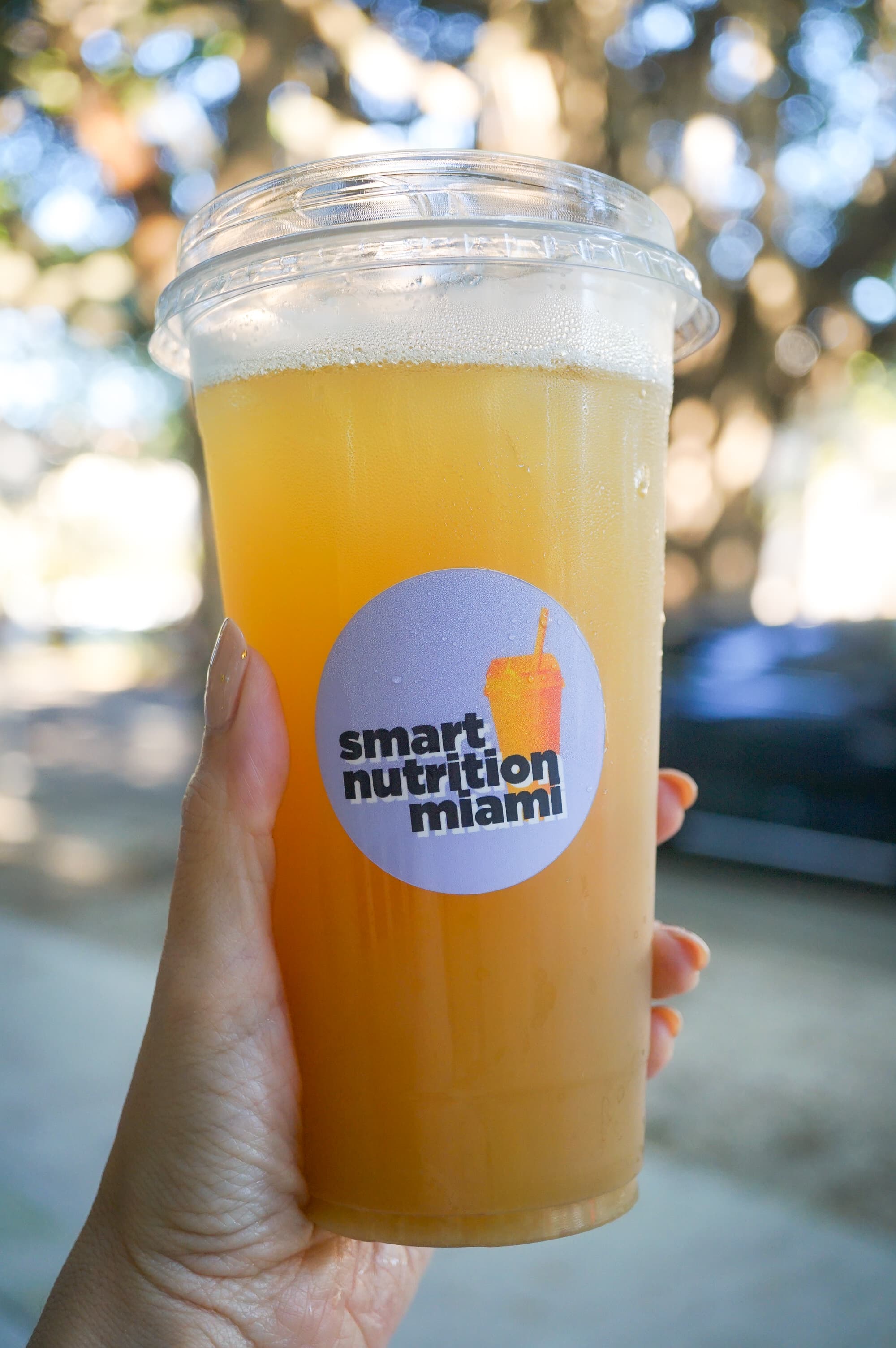 Hand holding a clear plastic cup of yellow juice with a Smart Nutrition Miami sticker.