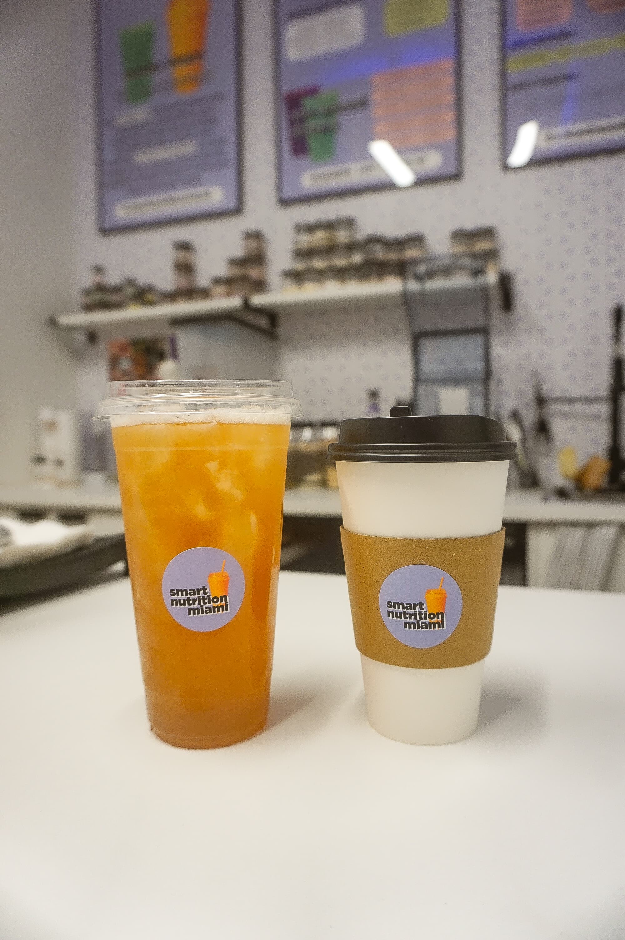 Iced orange tea and hot coffee in branded cups from Smart Nutrition Miami on counter.